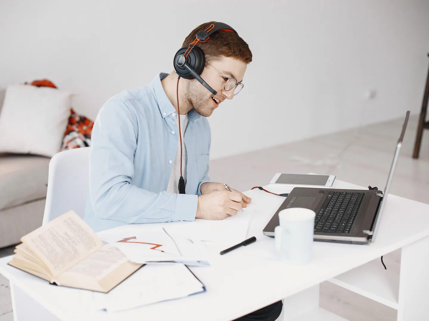 A man wearing a headset and taking notes beside a laptop in a home workspace.